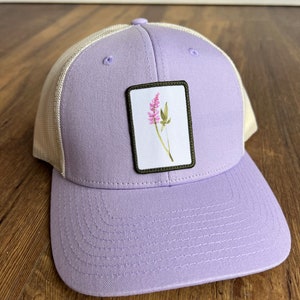 May include: A lavender trucker hat with a white mesh back and a patch featuring a pink flower with green leaves.