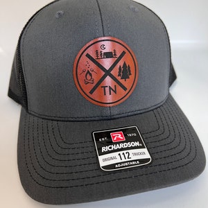 May include: Gray and black trucker hat with a brown leather patch featuring a campfire, tent, and trees with the text "TN" inside a circle. The hat has the text "Richardson" and "112" on the front.