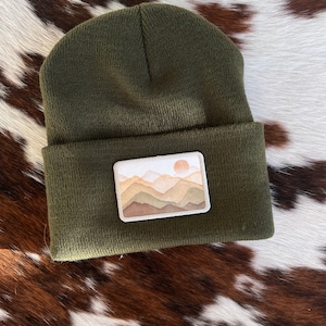 May include: A green beanie with a patch featuring a watercolor-style mountain range with a sun setting behind it.