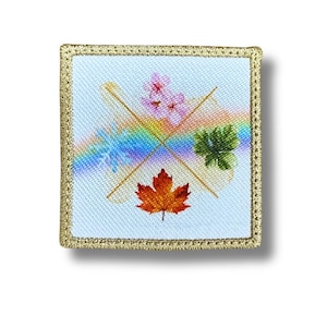 May include: Square patch with a light blue background and a gold border. The patch features a rainbow, a red maple leaf, pink flowers, and green leaves. Two gold lines cross the center of the patch.