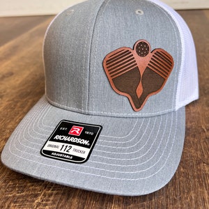 May include: Gray and white trucker hat with a brown leather patch of two pickleball paddles and a ball. The hat has the text "EST. 1970 RICHARDSON. ORIGINAL 112 TRUCKER ADJUSTABLE" on the back.