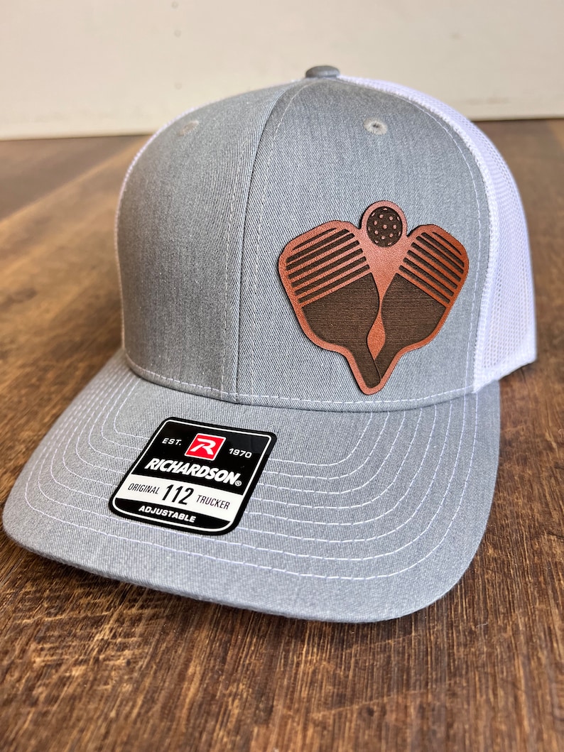 Pu&ograve; includere: Cappellino da camionista grigio e bianco con una toppa in pelle marrone di due racchette da pickleball e una palla. Il cappello ha il testo "EST. 1970 RICHARDSON. ORIGINAL 112 TRUCKER ADJUSTABLE" sul retro.