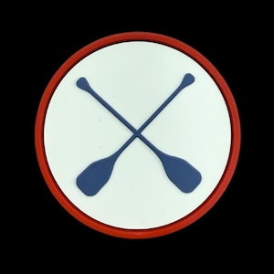 May include: A round, white emblem with a red border featuring two crossed blue oars. The oars are positioned diagonally across the center of the emblem, creating an 'X' shape. The design is set against a black background.