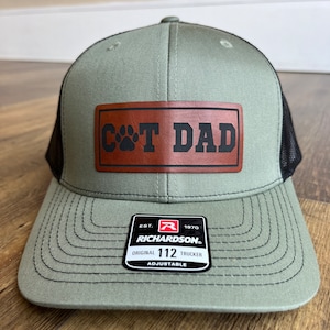 May include: A green and black trucker hat with a brown leather patch that says "CAT DAD" with a paw print. The hat is adjustable and has a Richardson 112 label.