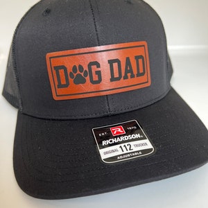 May include: Black trucker hat with a mesh back and a curved brim. The hat features a rectangular brown patch with the words "DOG DAD" and a paw print. The hat has an adjustable strap and a tag that reads "RICHARDSON 112 TRUCKER".