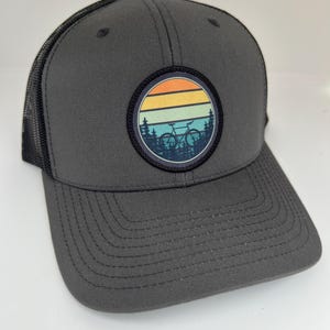 May include: A gray trucker hat with a black mesh back. The hat features a round patch with a vintage sunset design and a bicycle silhouette.