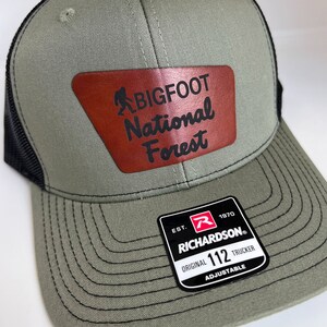 May include: A green and black trucker hat with a brown leather patch that says "BIGFOOT National Forest". The hat is adjustable and has a Richardson 112 label.