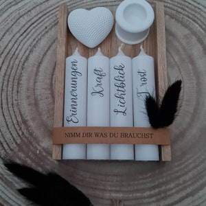 May include: Four white candles in a wooden box with the words "Erinnerungen", "Kraft", "Lichtblick", and "Trost" written on them. The box also has a small white heart and a white candle holder. The text "Nimm dir was du brauchst" is written on the box.