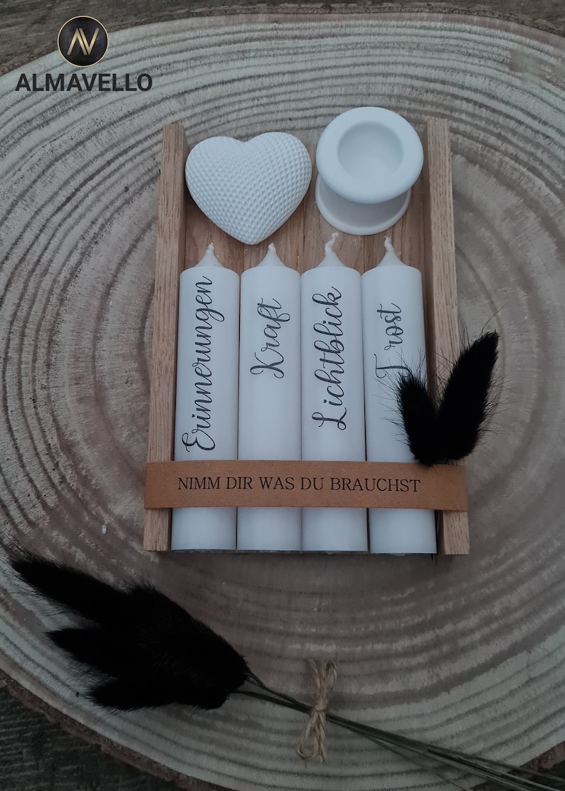 May include: Four white candles in a wooden box with the words "Erinnerungen", "Kraft", "Lichtblick", and "Trost" written on them. The box also has a small white heart and a white candle holder. The text "Nimm dir was du brauchst" is written on the box.