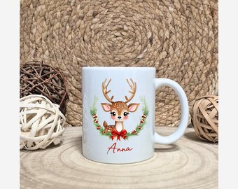 Könnte beinhalten: Weiße Keramiktasse mit einem Cartoon-Hirsch-Design, einschließlich Geweih, roter Schleife und grünem Kranz. Der Name "Anna" ist in roter Schrift geschrieben. Die Tasse hat einen weißen Griff.