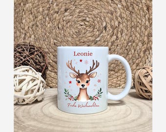 Könnte beinhalten: Weiße Keramik-Tasse mit einem Cartoon-Hirsch, roten Sternen und Schneeflocken. Die Tasse trägt den Namen "Leonie" in Rot und den Schriftzug "Frohe Weihnachten" in Rot. Die Tasse hat einen weißen Henkel und steht auf einer Holzoberfläche.