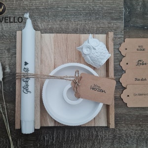 Könnte beinhalten: Eine weiße Kerze mit dem Text "Glückssicht" darauf, eine weiße Eulenfigur, ein weißer Kerzenhalter und vier braune Geschenkanhänger mit dem Text "Viel Glück", "Mit Liebe", "Für dich" und "Ein kleiner Gruß".