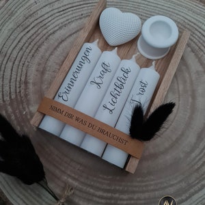 May include: A wooden box with four white candles and a white heart. The candles are labeled "Erinnerungen", "Kraft", "Lichtblick", and "Trost". The box has a brown leather strap with the text "Nimm dir was du brauchst".
