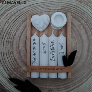 May include: Four white candles in a wooden box with the words "Erinnerungen", "Kraft", "Lichtblick", and "Trost" printed on them. A white heart and a small white dish are also in the box. The box has a brown leather strap with the text "Nimm dir was du brauchst" printed on it.