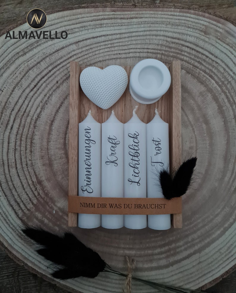 May include: Four white candles in a wooden box with the words "Erinnerungen", "Kraft", "Lichtblick", and "Trost" printed on them. A white heart and a small white dish are also in the box. The box has a brown leather strap with the text "Nimm dir was du brauchst" printed on it.