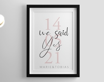 May include: A framed print with a black border, featuring the text "14 we said Yes 21" in a mix of pink and black fonts. The names "MARIE & TOBIAS" are at the bottom. Decorative spheres hang in the upper left.
