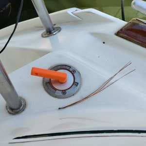 May include: A close-up of a white boat deck with a silver fuel cap labeled "Diesel" and an orange lever. The lever is in the "Off" position. A few brown pine needles are lying on the deck.