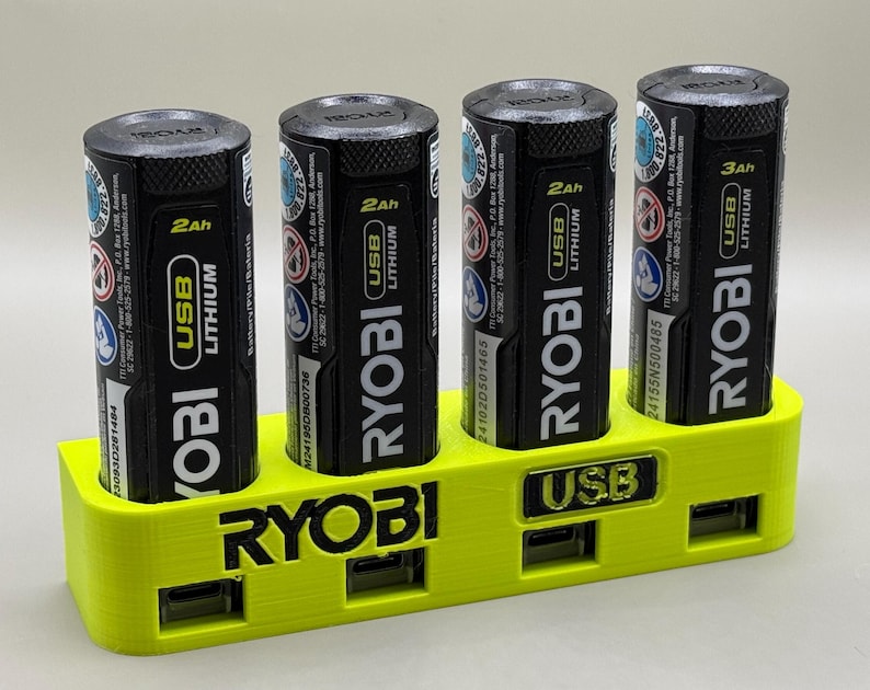 K&ouml;nnte beinhalten: Eine gelbe Ladestation aus Kunststoff f&uuml;r vier Ryobi USB-Lithium-Akkus. Die Station hat vier Steckpl&auml;tze f&uuml;r die Akkus und zwei USB-Anschl&uuml;sse an der Vorderseite.