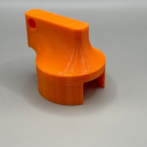 Puede incluir: Un objeto de plástico naranja impreso en 3D con un asa y un agujero en la parte superior. El objeto tiene una base redondeada y una abertura rectangular en el lateral.