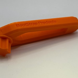 May include: Bright orange 3D-printed tool with the text "BootStrap Products" on the side. The tool has a handle with a cutout and a star-shaped end. The object is on a white surface.