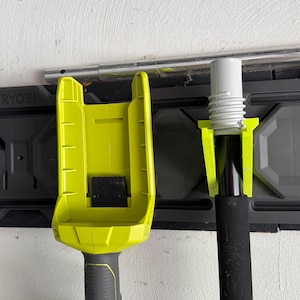 Könnte beinhalten: Ein gelb-schwarzer Ryobi-Werkzeughalter, montiert an einem schwarzen Wandorganizer. Der Halter ist für die Sicherung eines Werkzeugs mit schwarzem Griff konzipiert. Ein weißes und graues Werkzeug wird ebenfalls in einem gelben Halter gehalten.