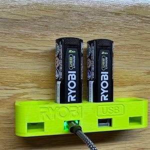 K&ouml;nnte beinhalten: Eine gr&uuml;ne Wandhalterung mit dem Text "RYOBI USB" darauf. Zwei schwarze RYOBI 2Ah USB Lithium-Akkus sind in die Wandhalterung gesteckt.