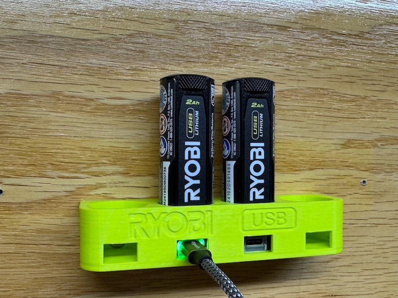 K&ouml;nnte beinhalten: Eine gr&uuml;ne Wandhalterung mit dem Text "RYOBI USB" darauf. Zwei schwarze RYOBI 2Ah USB Lithium-Akkus sind in die Wandhalterung gesteckt.
