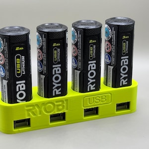K&ouml;nnte beinhalten: Ein gelber 3D-gedruckter Batterieladeger&auml;t mit vier Steckpl&auml;tzen zum Aufnehmen von Batterien. Das Ladeger&auml;t hat den Schriftzug "RYOBI" auf der Vorderseite und den Schriftzug "USB" auf der Seite. Das Ladeger&auml;t h&auml;lt vier schwarze RYOBI-Batterien mit dem Aufdruck "RYOBI USB LITHIUM" auf ihnen.