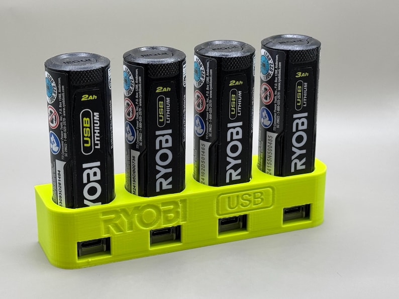 K&ouml;nnte beinhalten: Ein gelber 3D-gedruckter Batterieladeger&auml;t mit vier Steckpl&auml;tzen zum Aufnehmen von Batterien. Das Ladeger&auml;t hat den Schriftzug "RYOBI" auf der Vorderseite und den Schriftzug "USB" auf der Seite. Das Ladeger&auml;t h&auml;lt vier schwarze RYOBI-Batterien mit dem Aufdruck "RYOBI USB LITHIUM" auf ihnen.