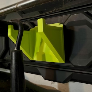 Può includere: Primo piano di un porta attrezzi verde lime attaccato a un sistema di stoccaggio a parete nero. Un attrezzo nero è parzialmente inserito nel supporto. Il supporto è in plastica e ha un design geometrico.