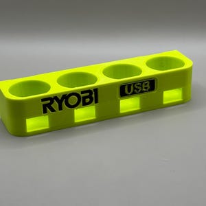 K&ouml;nnte beinhalten: Ein leuchtend gelber Kunststoffhalter mit vier Steckpl&auml;tzen f&uuml;r Batterien. Der Halter tr&auml;gt den Schriftzug "RYOBI" und einen kleineren Steckplatz mit der Aufschrift "USB".