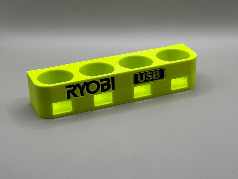 K&ouml;nnte beinhalten: Ein leuchtend gelber Kunststoffhalter mit vier Steckpl&auml;tzen f&uuml;r Batterien. Der Halter tr&auml;gt den Schriftzug "RYOBI" und einen kleineren Steckplatz mit der Aufschrift "USB".