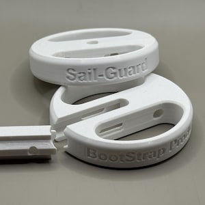 May include: White plastic Sail-Guard and BootStrap Pro boat accessories. The image shows two circular pieces and a rectangular piece. The text "Sail-Guard" and "BootStrap Pro" are embossed on the plastic. The pieces are designed to be used together.