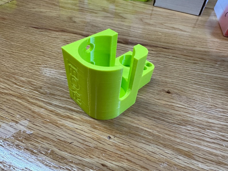 Peut inclure: Un support mural imprim&eacute; en 3D de couleur vert vif avec le mot "BOOST" imprim&eacute; sur le c&ocirc;t&eacute;. Le support a une ouverture circulaire et une fente pour maintenir un objet.