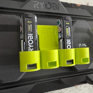 K&ouml;nnte beinhalten: Eine gr&uuml;ne Wandhalterung mit drei Steckpl&auml;tzen zum Aufbewahren von Ryobi 2Ah Lithium-Ionen-Akkus. Die Akkus haben einen USB-Anschluss und sind mit "Ryobi" und "Lithium" beschriftet.