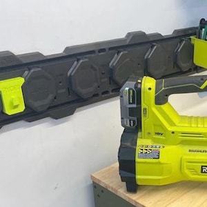 Può includere: Un soffiatore per foglie a batteria Ryobi verde lime e nero con batteria da 18V, posizionato vicino a un sistema di binari a parete nero con ganci accessori verde lime e un contenitore verde lime per organizzare gli attrezzi.