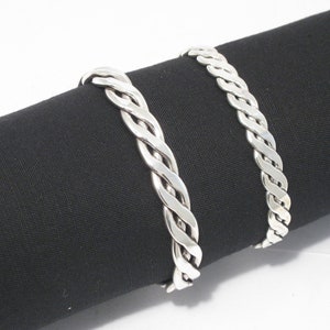 May include: Two silver braided bracelets, one slightly larger than the other, are displayed on a black fabric background.