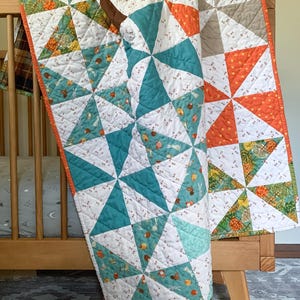 May include: A handmade quilt with a pinwheel design in teal, orange, and white. The quilt is draped over a wooden crib, showcasing its intricate patchwork and warm color palette. The quilt features a variety of fabrics with small animal and floral patterns.
