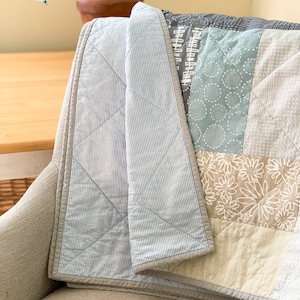 May include: A blue and white striped quilt with a patchwork design featuring various patterns in shades of grey, blue, and beige. The quilt is folded and draped over a chair.
