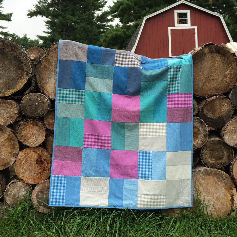 May include: A patchwork quilt made from different shades of blue fabric with pink, white, and grey squares. The quilt is hanging on a pile of logs in front of a red barn.