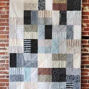May include: A patchwork quilt with a white background and various black, white, and grey patterned squares. The quilt is hanging on a brick wall.