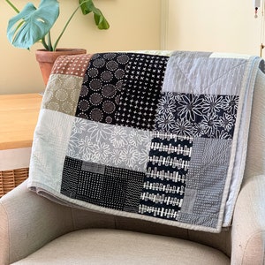 May include: A patchwork quilt featuring a range of black and white patterns, including geometric shapes, flowers, and dots. The quilt is folded and draped over a chair.