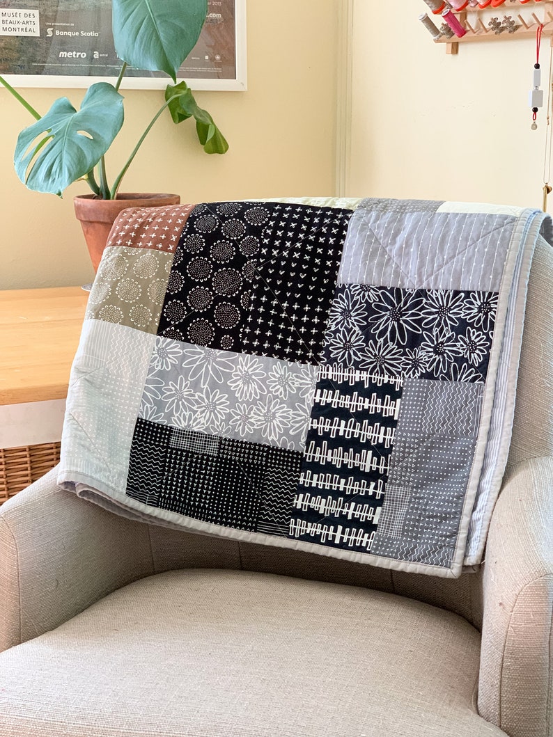 May include: A patchwork quilt featuring a range of black and white patterns, including geometric shapes, flowers, and dots. The quilt is folded and draped over a chair.