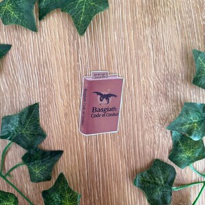 May include: A sticker featuring a maroon book with the title "Basgiath Code of Conduct" and a black dragon illustration. The sticker has the text "Spich Eye Section One" on the top and is surrounded by green ivy.