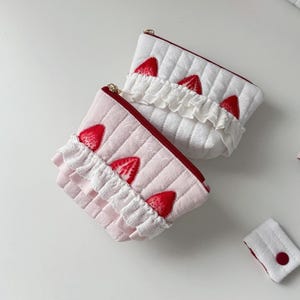 May include: Two quilted pouches, one white and one light pink, featuring red strawberry appliques and white ruffled lace trim. Each pouch has a red zipper closure along the top edge. A small, rectangular white quilted tab with a red button is included.