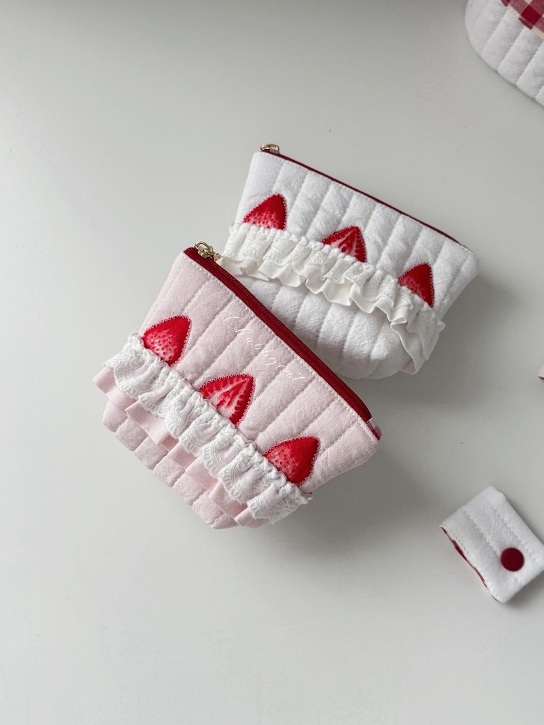 May include: Two quilted pouches, one white and one light pink, featuring red strawberry appliques and white ruffled lace trim. Each pouch has a red zipper closure along the top edge. A small, rectangular white quilted tab with a red button is included.