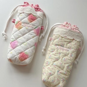 May include: Two quilted drawstring pouches with white lace trim and pink gingham fabric at the top. The left pouch features a white background with pink strawberry patterns, while the right pouch has a pale yellow background with small floral prints.