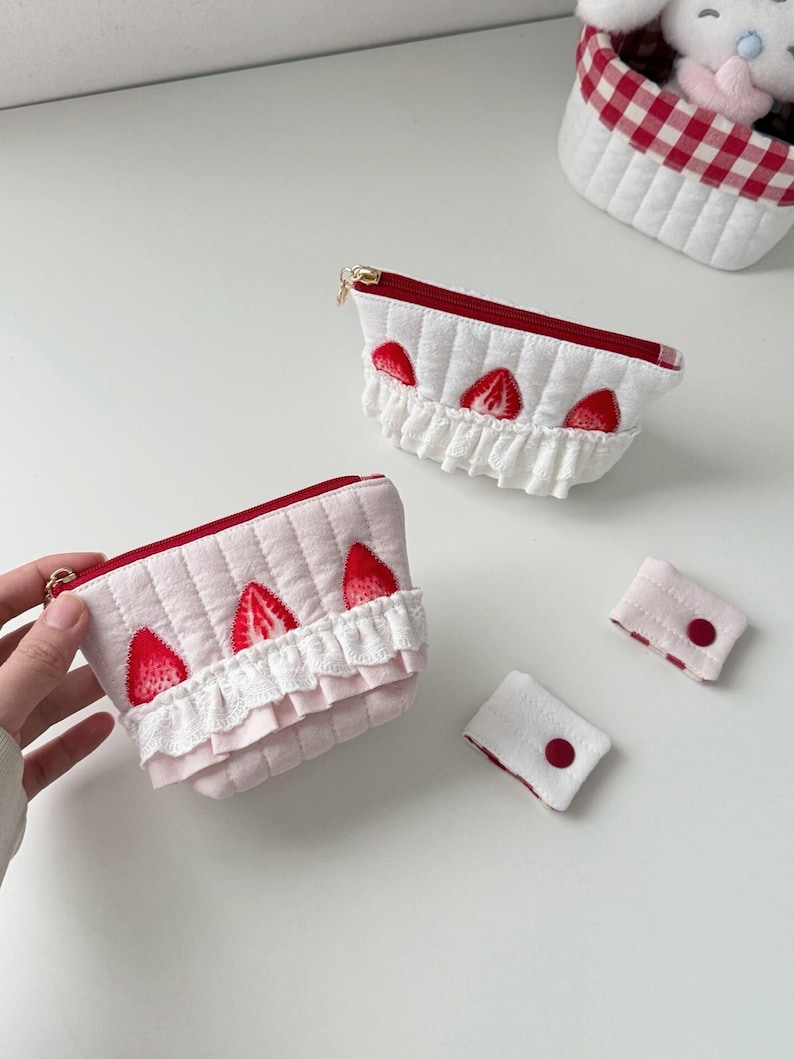 May include: Two quilted pouches featuring red strawberry appliques and white ruffled lace trim along the front. One pouch is white with a red zipper, and the other is light pink with a red zipper. Two small rectangular fabric tabs with red buttons are included.