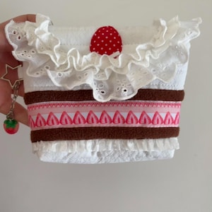 May include: A white fabric pouch featuring a ruffled eyelet trim at the top and a red polka-dot accent. The center includes brown fabric bands and a pink ribbon with a repeating strawberry pattern. A star-shaped charm and a strawberry charm hang from the side.