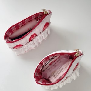 May include: Two quilted pouches featuring a white exterior with red strawberry prints and white ruffled trim. The interiors are lined with red and white gingham fabric. Each pouch has a gold zipper closure and contains various small personal items.
